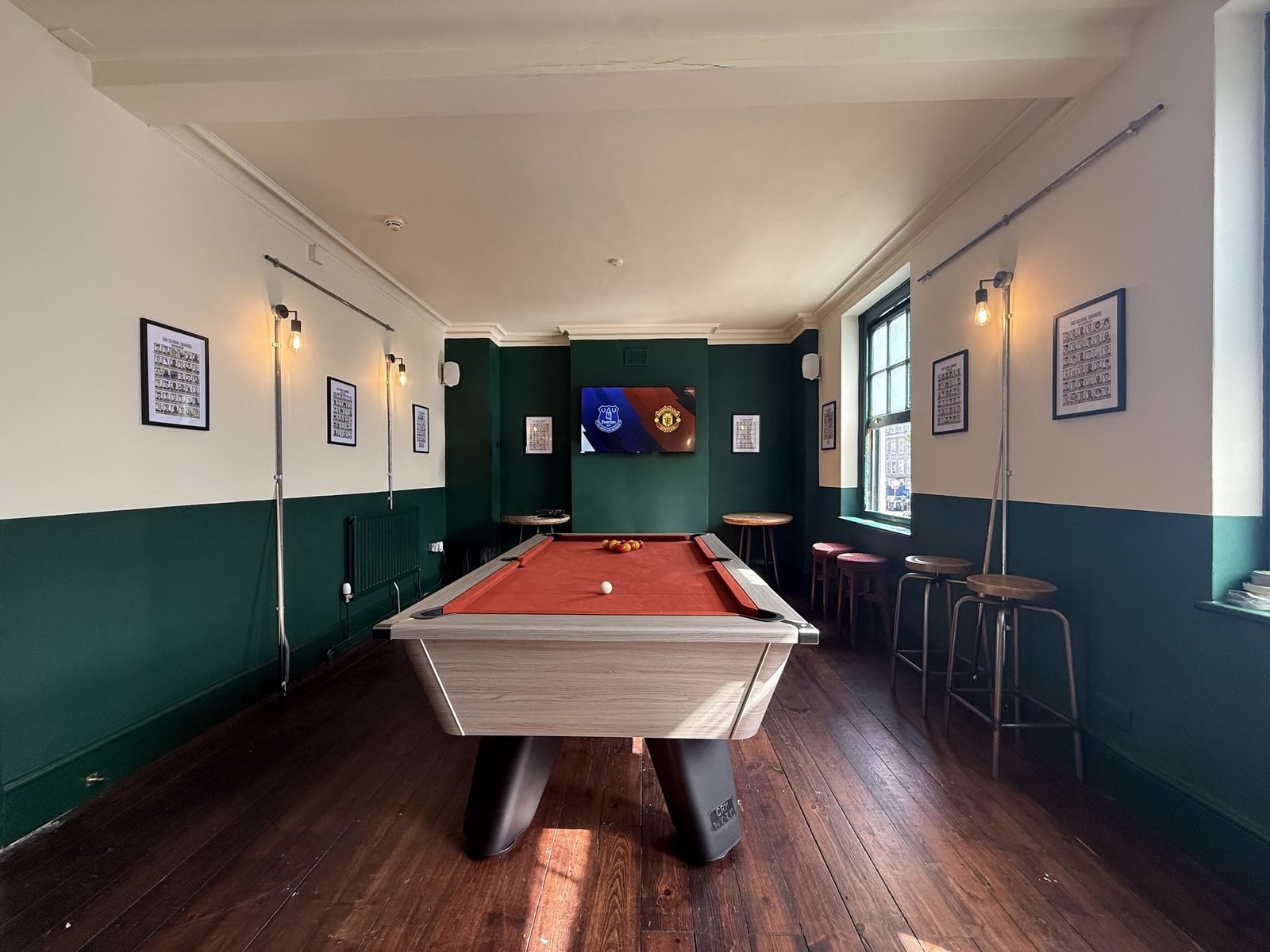The pool room: the pool table with its red cloth and the balls racked, green-panelled walls, framed prints, wall lamps and football on the TV at the far end